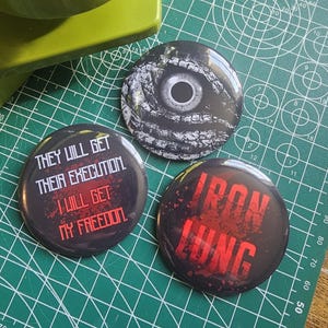 May include: Three circular badges with dark designs. One shows an eye graphic, another reads "THEY WILL GET THEIR EXECUTION. I WILL GET MY FREEDOM.", and the last says "IRON LUNG" in red.