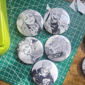 Pode incluir: Um conjunto de cinco botões redondos em preto e branco com ilustrações de personagens de anime. Cada botão apresenta um personagem diferente. Os botões estão dispostos sobre uma base de corte verde com medidas.