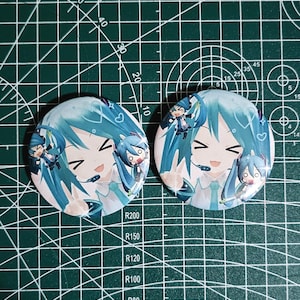 May include: Two circular white badges featuring a smiling anime character with long, turquoise hair and a headset. The character is surrounded by hearts and smaller versions of itself. The badges are on a green cutting mat.