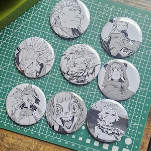 May include: A collection of nine round, black and white manga-style character button pins. The pins feature various detailed illustrations of anime-style characters. The pins are arranged on a green cutting mat.