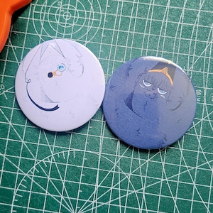 May include: Two circular badges featuring cartoon illustrations. One is white with blue and gold details, the other is blue with yellow and black. They rest on a green cutting mat.