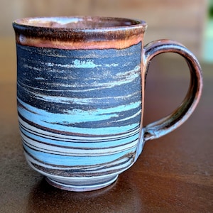 May include: Handmade ceramic mug with a unique, textured design. The mug features horizontal bands of black, blue, and white, with a brown rim and handle. This stoneware mug is perfect for coffee, tea, or any hot beverage.