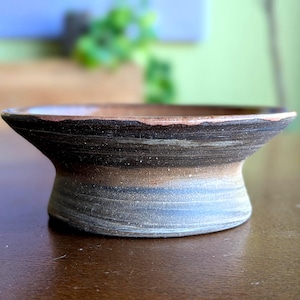 May include: A handmade ceramic bowl with a flared base and a wide, shallow top. The bowl features a layered design with shades of brown, black, and blue. The rim has a reddish-brown hue. The bowl is likely used for serving or display.