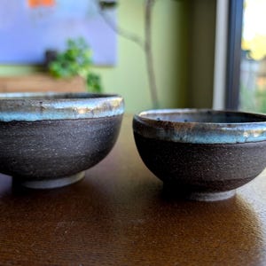 May include: Two handmade ceramic bowls. The bowls are dark brown with a textured surface and a light blue glaze around the rim. The bowls are of different sizes and sit on a wooden surface.
