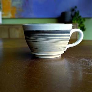 Op de afbeelding: Een keramische koffiekop met een witte oor en rand. De kop heeft een horizontaal gestreept patroon in grijstinten en beige. De kop staat op een donkerbruin houten oppervlak.