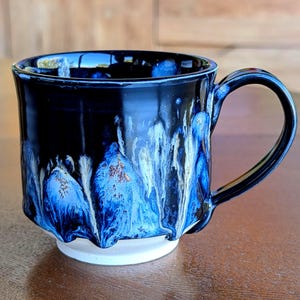 Op de afbeelding: Een handgemaakte keramische mok met een zwarte buitenkant en een opvallend blauw-wit glazuurontwerp. De mok heeft een afgerond oor en staat op een witte basis. De binnenkant is zwart, voor warme dranken.