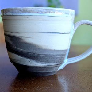 Op de afbeelding: Een handgemaakte keramische mok met een uniek gemarmerd ontwerp. De mok heeft een wit en grijs wervelpatroon, een afgeronde handgreep en een licht uitlopende rand. Geschikt voor koffie, thee of andere dranken.