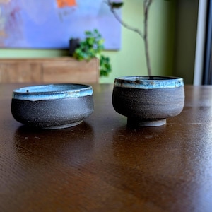 May include: Two small, handmade ceramic bowls. The bowls are dark brown with a textured surface and a light blue rim. One bowl is low and wide, while the other has a small footed base. They sit on a wooden table.