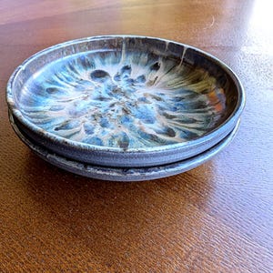 May include: Two ceramic plates with a dark rim and a blue and white marbled glaze. The plates are stacked, highlighting the unique patterns and textures of the handmade pottery. The plates are on a wooden surface.