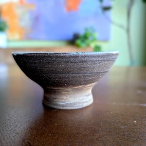 May include: A handmade, dark brown ceramic bowl with a light-coloured base. The bowl has a wide, shallow shape with a textured surface and a slightly flared rim. The bowl is sitting on a brown surface.