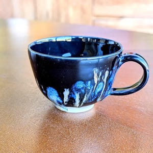May include: A black ceramic teacup with a rounded base and a handle. The cup's exterior has a dark, glossy finish with blue and white dripped glaze patterns. The cup sits on a light-coloured surface.