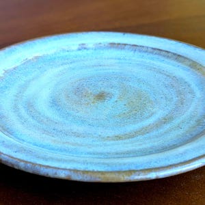 May include: A light blue ceramic plate with a swirling pattern. The plate has a white glaze with a subtle blue tint and a brown rim.