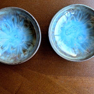May include: Two ceramic bowls with a distinctive glaze. The bowls are round with a raised rim. The interior has a vibrant blue and turquoise colour with a textured, flowing pattern. The exterior has a darker, earthy tone, creating a contrast.