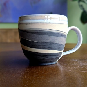 Op de afbeelding: Een keramische mok met een wit handvat en een cilindrisch lichaam. De mok heeft een gelaagd ontwerp van zwarte, grijze en beige horizontale banden. De mok staat op een bruin houten oppervlak.