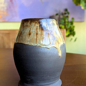 Op de afbeelding: Een kleine, zwarte keramische vaas met een bruine en groene glazuur. De glazuur heeft een druppel effect op de bovenrand van de vaas.