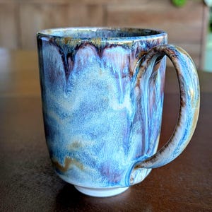 May include: A ceramic mug with a distinctive glaze in blue, brown, and gold hues. The glaze runs down the sides, producing a textured appearance. The mug has a white base and a large handle, suitable for tea or coffee.