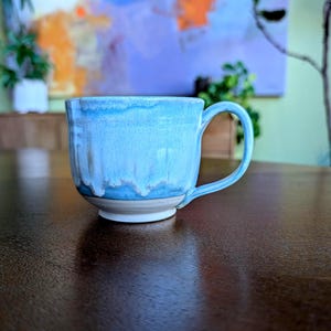 May include: A ceramic mug with a light blue glaze and a white base. The mug has a rounded shape with a handle and is on a dark brown wooden table. The mug's glaze has a dripped effect, creating a unique texture. The mug is likely handmade.