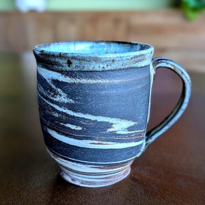 May include: Handmade ceramic mug with a unique marbled design. The mug features a dark blue exterior with white and brown streaks, a light blue rim, and a curved handle. This stoneware mug is perfect for coffee, tea, or any warm beverage.
