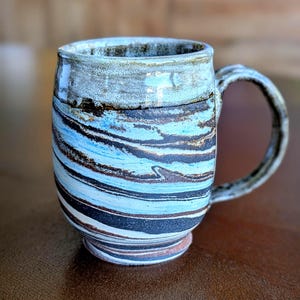 May include: Handmade ceramic mug with a unique swirl design in shades of blue, brown, and black. The mug has a sturdy handle and a textured rim. The mug is a perfect size for coffee or tea.