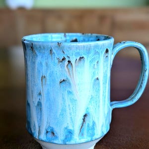 May include: A ceramic mug with a light blue glaze and a white base. The mug has a handle and a cylindrical shape. The glaze has a dripped effect with brown and white accents. The mug is suitable for hot or cold drinks.