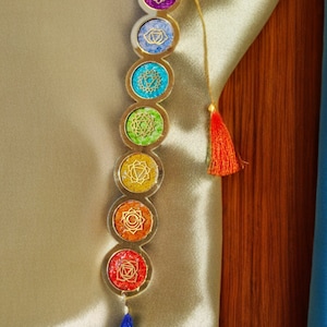 Puede incluir: Un colgante de chakra vertical con siete diseños circulares coloridos. Cada círculo presenta un color y un símbolo diferente, con un borde dorado. El colgante tiene una cuerda dorada y una borla roja y azul.