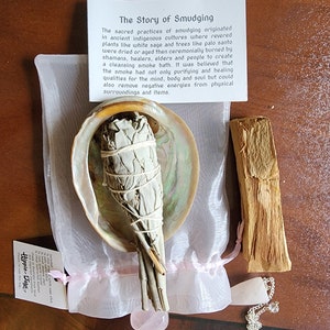 Puede incluir: Un palo de salvia blanca atado con cuerda, descansando en una concha de abulón blanca. También se incluye un palo de palo santo y un cristal de cuarzo rosa. El texto "The Story of Smudging" está impreso en una tarjeta blanca.