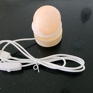 Pode incluir: Uma pequena lâmpada de sal redonda e laranja com uma base de madeira e um cabo branco com um interruptor. A lâmpada está ligada e acesa.