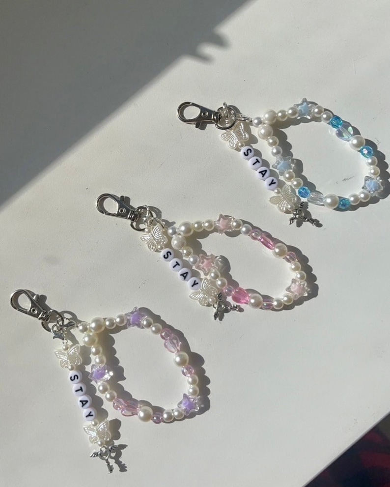 May include: Three beaded keychains with silver clasps. Each keychain features a white pearl and colourful bead design with a silver butterfly charm and the word "STAY" spelled out in white letters.