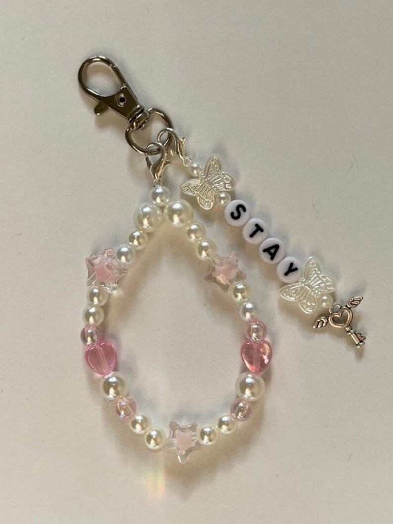 May include: A white pearl and pink heart charm keyring with a silver key charm, a butterfly charm, and the word "STAY" spelled out in white beads.
