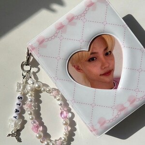 May include: A white photo album with a pink and white heart-shaped cutout featuring a photo of a young man. The album has a pink and white patterned design and comes with a pearl and pink bead charm bracelet with a silver charm that says "STAY".