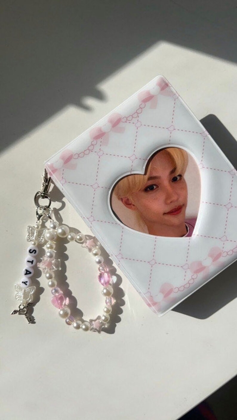 May include: A white photo album with a pink and white heart-shaped cutout featuring a photo of a young man. The album has a pink and white patterned design and comes with a pearl and pink bead charm bracelet with a silver charm that says "STAY".