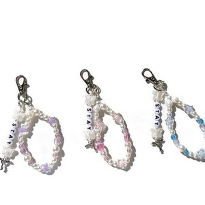 May include: Three beaded keychains with silver clasps. Each keychain features a double strand of white pearls with colourful accents and a charm of a butterfly. The charms are labelled "STAY" in white letters.