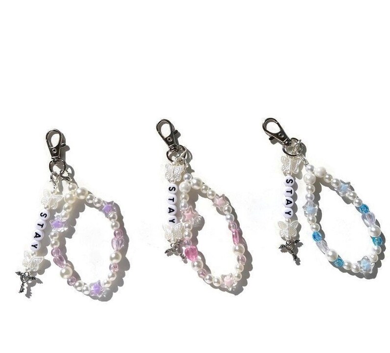 May include: Three beaded keychains with silver clasps. Each keychain features a double strand of white pearls with colourful accents and a charm of a butterfly. The charms are labelled "STAY" in white letters.