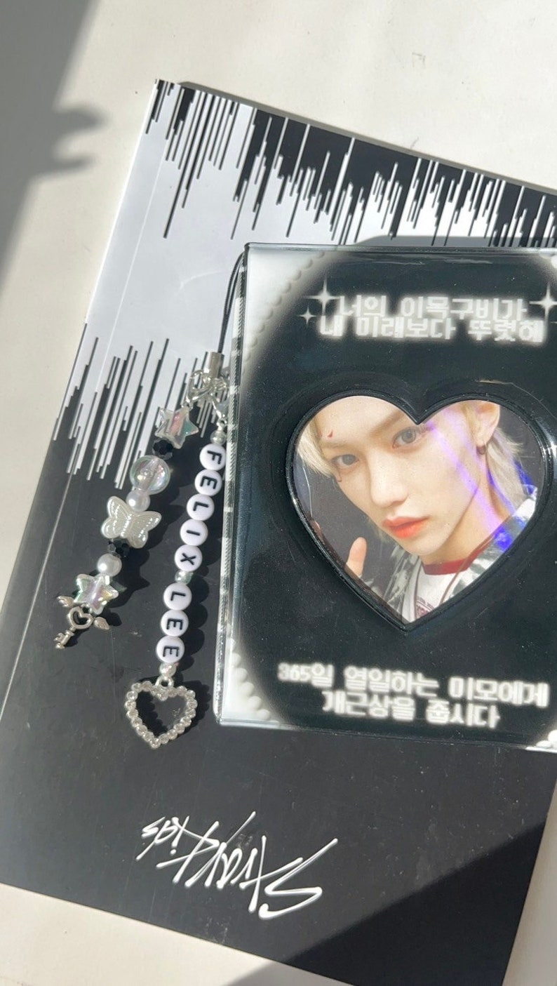May include: A black and white photo card with a heart-shaped frame featuring a portrait of a young man with blonde hair. The card has the text "너의 이목구비가 미래보다 뚜렷해" and "365일 열일하는 미모에게 개근상을 줍시다" in Korean. A black and white beaded charm with the name "FELIX LEE" is attached to the card.