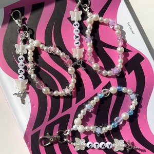 May include: Three white pearl beaded keychains with silver charms and the word "STAY" spelled out in white letters. Each keychain has a different coloured accent bead: pink, blue, and purple.