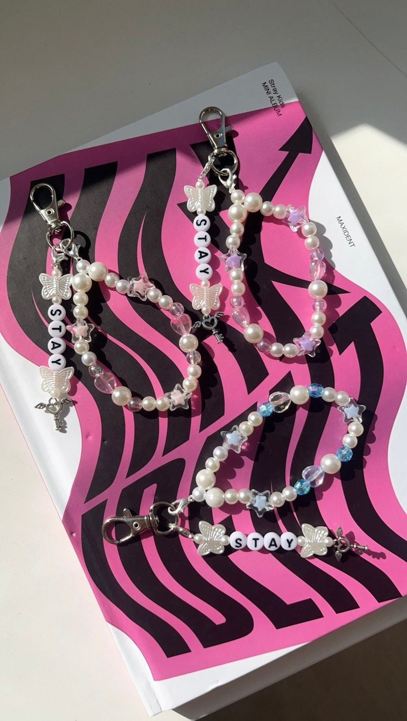 May include: Three white pearl beaded keychains with silver charms and the word "STAY" spelled out in white letters. Each keychain has a different coloured accent bead: pink, blue, and purple.