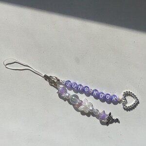 May include: Purple and white beaded phone charm with the name "JUNGKOOK" spelled out in white letters. The charm also features a silver butterfly, a silver key, and a silver heart.