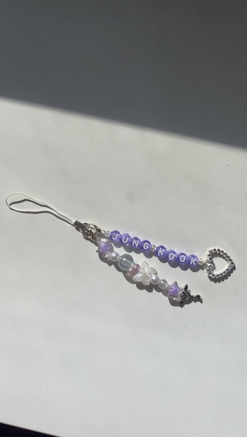 May include: Purple and white beaded phone charm with the name "JUNGKOOK" spelled out in white letters. The charm also features a silver butterfly, a silver key, and a silver heart.