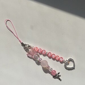 May include: A pink phone charm with a silver heart charm, pink beads, and the word "LEEKNOW" spelled out in white letters.