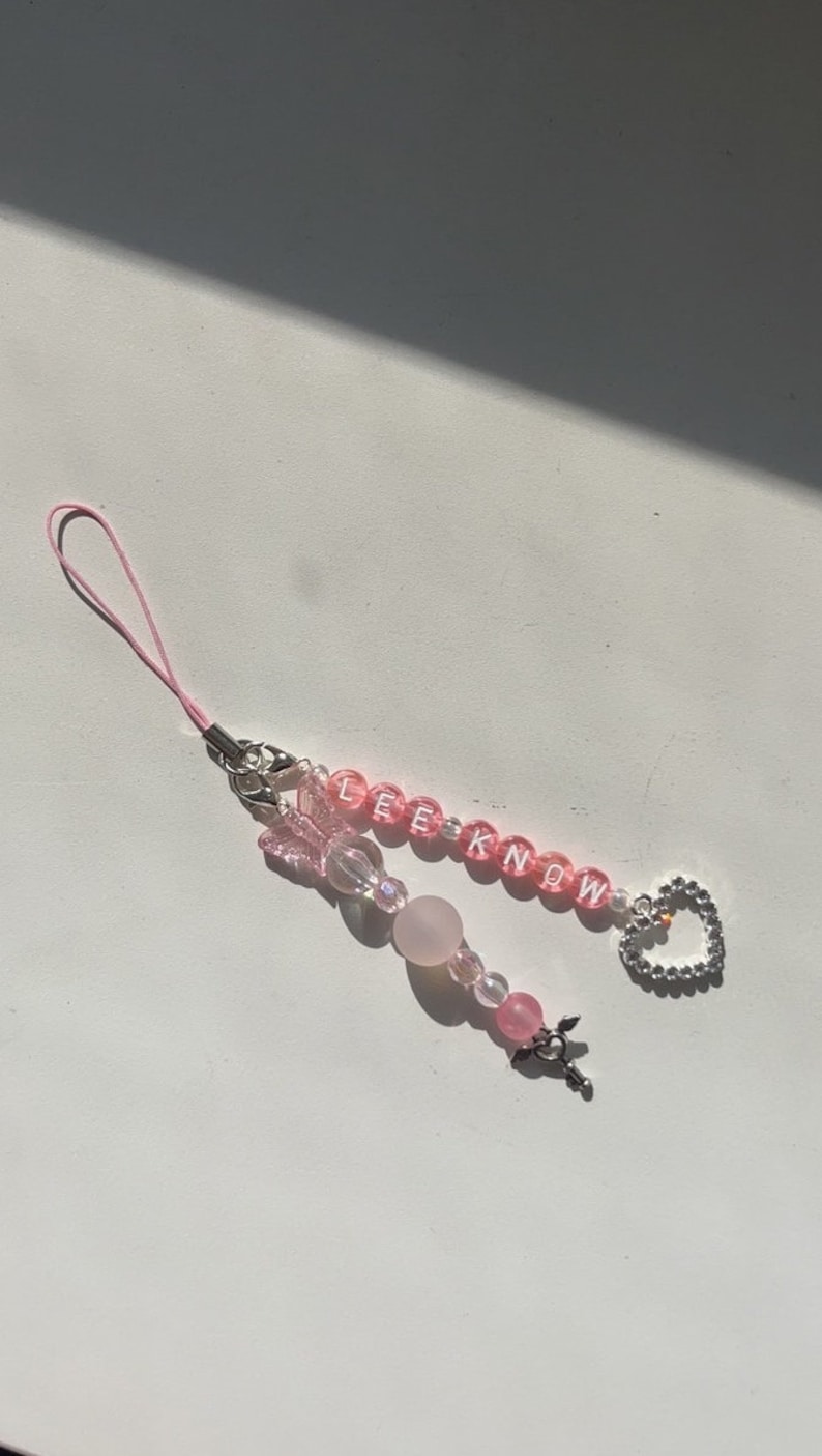 May include: A pink phone charm with a silver heart charm, pink beads, and the word "LEEKNOW" spelled out in white letters.