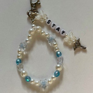 May include: A white pearl and blue crystal beaded keychain with a silver charm, a butterfly charm, and the word "STAY" spelled out in white letters.