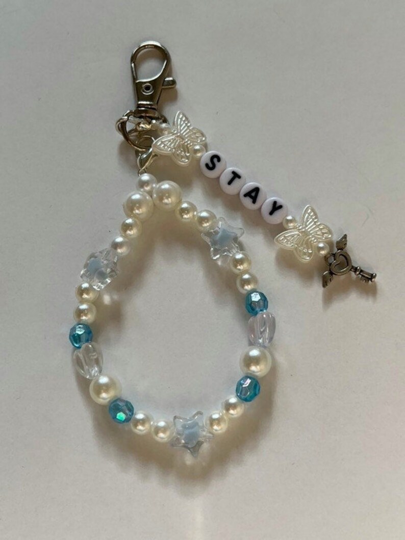 May include: A white pearl and blue crystal beaded keychain with a silver charm, a butterfly charm, and the word "STAY" spelled out in white letters.