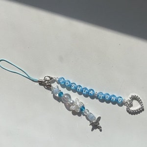 May include: A blue and white beaded phone charm with the name "Joshua Hong" spelled out in white letters. The charm also features a silver butterfly charm and a silver heart charm.