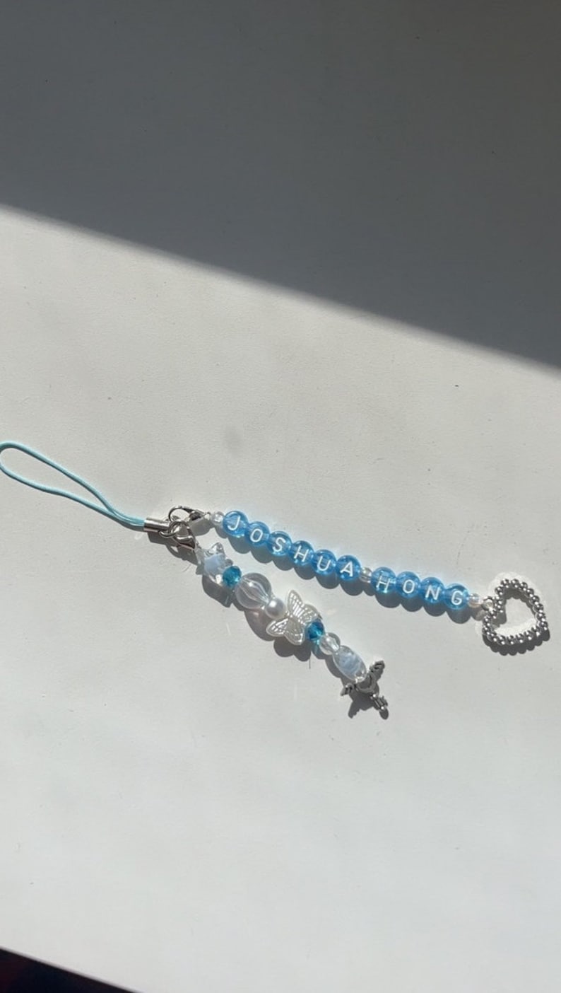 May include: A blue and white beaded phone charm with the name "Joshua Hong" spelled out in white letters. The charm also features a silver butterfly charm and a silver heart charm.
