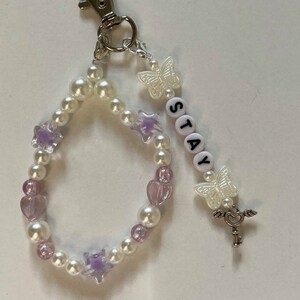 May include: A white and purple beaded keychain with a silver key charm, a butterfly charm, and the word "STAY" spelled out in white beads.