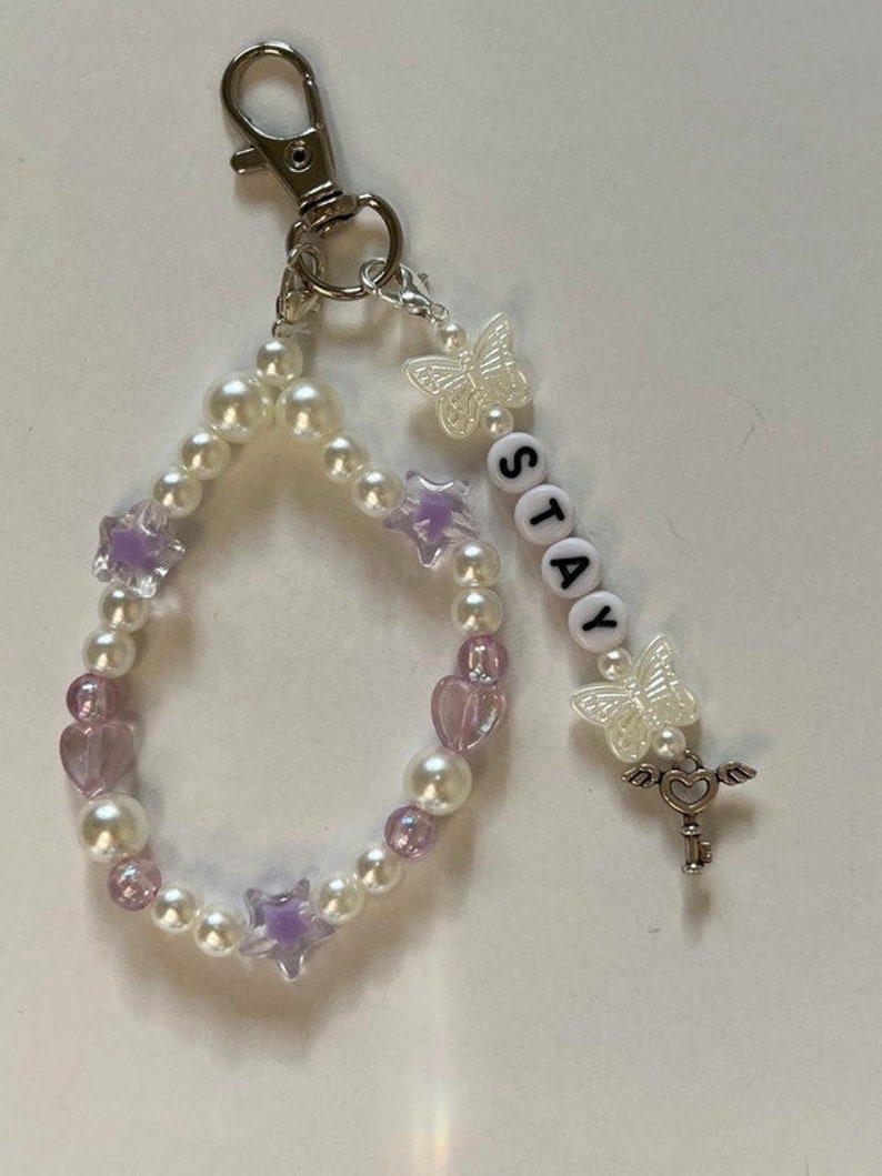May include: A white and purple beaded keychain with a silver key charm, a butterfly charm, and the word "STAY" spelled out in white beads.