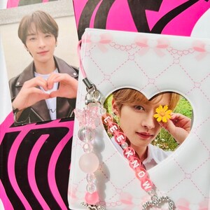 May include: A white and pink heart-shaped photo card with a picture of a young man with a flower behind his eye. The card is attached to a pink and white beaded keychain with the word "LEE KNOW" spelled out in pink beads.