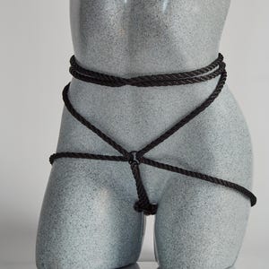 May include: A black rope harness with multiple straps and a metal ring. The harness is tied around a mannequin torso.