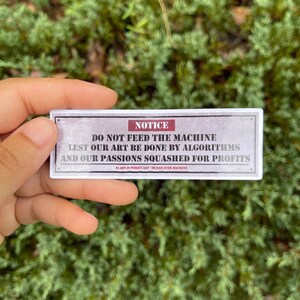 A rectangle die cut sticker of a warning sign with the message NOTICE, DO NOT FEED THE MACHINE, LEST OUR ART BE DONE BY ALGORITHMS AND OUR PASSIONS SQUASHED FOR PROFITS.