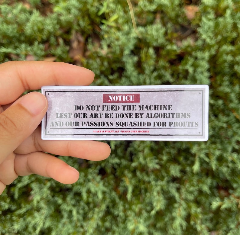 A rectangle die cut sticker of a warning sign with the message NOTICE, DO NOT FEED THE MACHINE, LEST OUR ART BE DONE BY ALGORITHMS AND OUR PASSIONS SQUASHED FOR PROFITS.